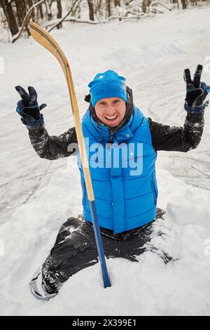 Young man poses standing on knees in snowdrift with hockey stick and shows victory. Stock Photo