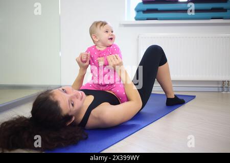 Happy kid sitting in her mothers belly lying on her back in the gym Stock Photo