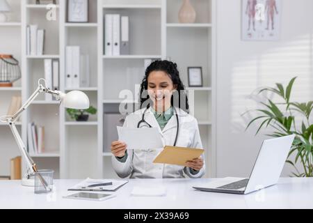 A cheerful Hispanic female doctor, in a white coat, reads an exciting letter or notification in a bright, modern medical office. Stock Photo