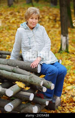 Elderly women in white poses on logs in autumn park with fallen leaves Stock Photo