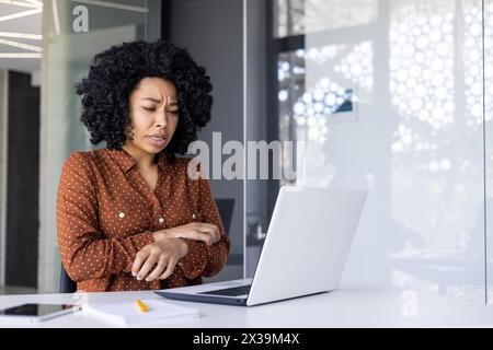 African American woman in office feeling itchy and distracted while working on her laptop, possibly experiencing an allergic reaction. Stock Photo