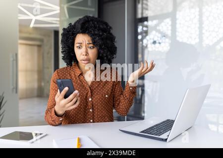 African American businesswoman looks shocked and confused while holding her smartphone in a modern office setting, reacting to unexpected news. Stock Photo