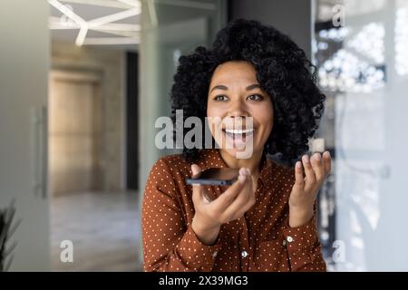Exuberant young woman with curly hair, dressed in a polka dot shirt, using voice commands on her smartphone in a modern office. Expressing joy and excitement. Stock Photo