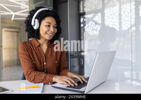 An African American young woman wearing a polka dot shirt and headphones, smiles as she works on her laptop in a modern office setting. Stock Photo