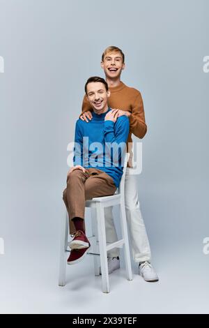 Two men, a loving gay couple in casual attire, sit on a stool posing for the camera against a gray backdrop. Stock Photo