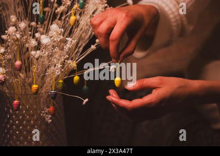 A woman hangs a yellow decorative Easter egg on a bouquet of dry white flowers in the interior. Festive home decor. Woman's hand holding toy pendant. Stock Photo