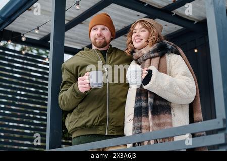 Low angle view medium shot of young Caucasian man and woman in love holding cups with hot drink standing on porch on winter day Stock Photo