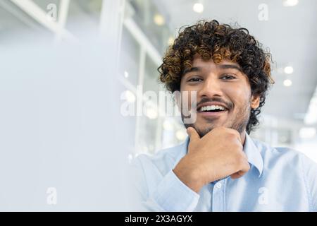 A cheerful young man with curly hair, wearing a light blue shirt, poses with a confident smile in a well-lit modern office setting, exuding positivity and professional confidence. Stock Photo