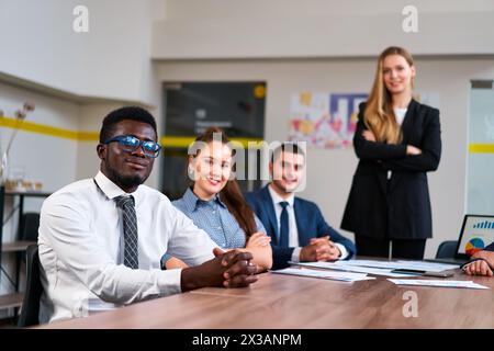 Multiethnic business team sits at meeting table in modern office. African, Caucasian, Arabic execs discuss strategy. Young woman leads diverse group Stock Photo