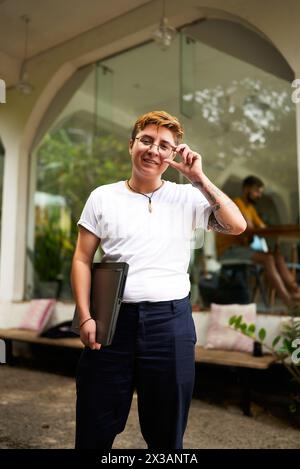 Transgender professional stands confidently in an office with laptop. Smiling, glasses-adjusting pose reflects inclusive, modern work environment Stock Photo