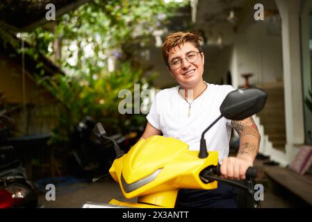 Smiling transgender individual on yellow scooter enjoys tropical setting. Person ready for ride, embracing freedom, warm climate. Confident pose with Stock Photo