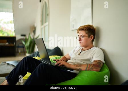 Transgender pro works on laptop from cozy bean bag in modern coworking space. Focused on screen, person enjoys flexible working environment. Inclusive Stock Photo