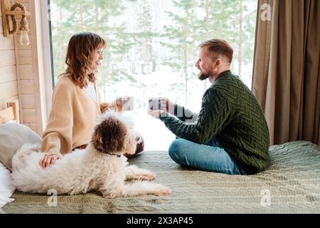 Modern Caucasian man and woman in love wearing casual clothes sitting on bed with their dog on it, drinking tea and talking Stock Photo
