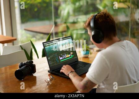 Pro videographer with headphones edits travel on laptop at coworking space, mirrorless camera beside. Creative freelancer works on video project Stock Photo