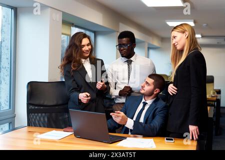 Multiethnic business team engages in lively discussion around laptop in modern office setting, with smiling woman standing, African man with papers Stock Photo