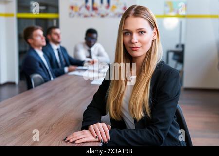 Confident blonde woman sits at boardroom table in modern office, team of male colleagues in background. Professional business team during meeting Stock Photo