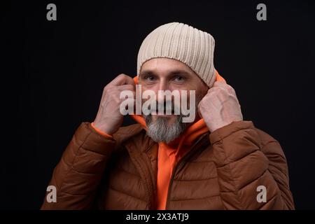A man in a brown jacket and orange hoodie is smiling and adjusting his hat. Concept of warmth and comfort, as the man is dressed for cold weather and Stock Photo