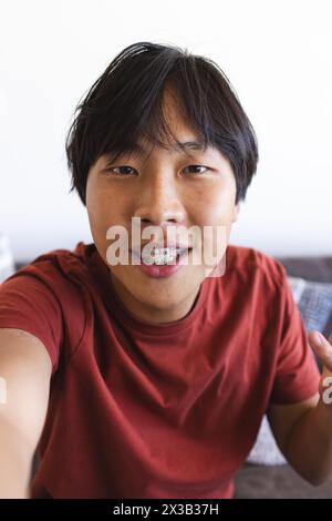 Asian teen boy with braces takes a selfie and video calls at home in a red shirt Stock Photo