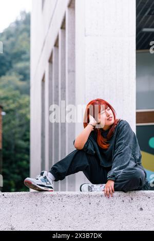 Stylish Korean woman with red hair sitting casually on concrete ledge Stock Photo
