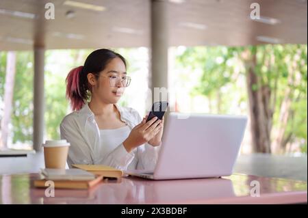 A young positive Asian female college student using her smartphone to check messages while sitting at a table in a campus cafeteria or canteen. Stock Photo