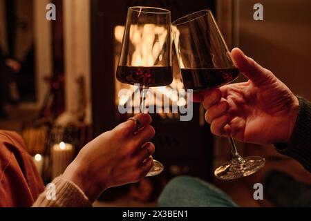 Hands of unrecognizable couple sitting in front of fireplace clinking glasses with wine while having date at home in evening Stock Photo