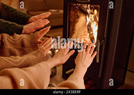 Unrecognizable family of three wearing sweaters sitting in front of fireplace at home warming their hands, copy space Stock Photo