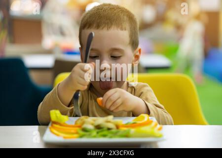 A funny cute three year old toddler boy eats fresh fruit while sitting at a table in a public place. Healthy food for children. Fruit plate. Focus on Stock Photo