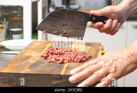 Cook chopping raw beef with a rustic kitchen cleaver on a wooden butcher block to make tartare, ground meat or minced meat, copy space, selected focus Stock Photo