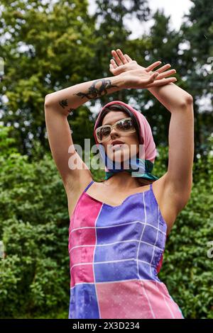 A beautiful young woman in a colorful dress joyfully raises her hands, enjoying the summer breeze in a natural setting. Stock Photo