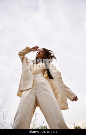 A young woman exudes elegance in a white suit and black tie, embracing the summer breeze in a serene field. Stock Photo