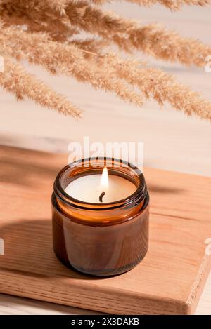 Cozy burning candle in amber glass jar, winter hygge home interior decor on wooden tray with dry reed or pampas grass bouquet. Scented wax eco candle. Stock Photo