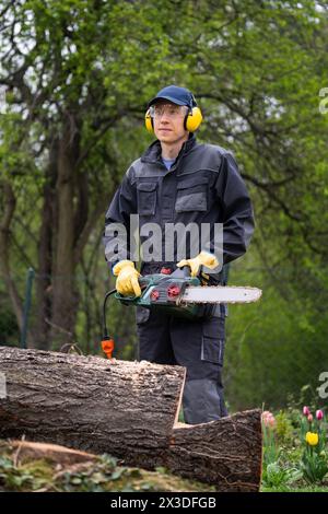 A man in uniform cuts an old tree in the yard with an electric saw. Stock Photo