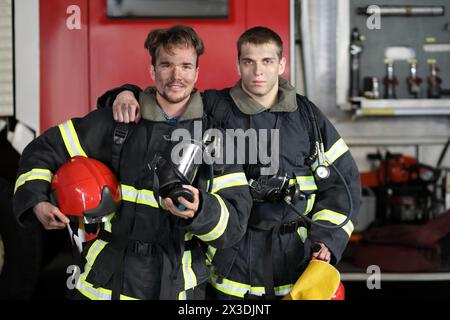 Two firefighters in protective suits standing against fire engine, one hugs other by shoulder Stock Photo