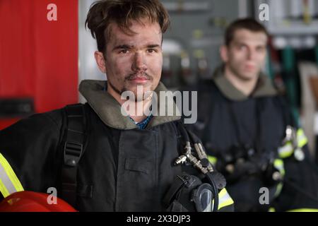 Two firefighters in protective suits with tool in hand without helmet against fire engine Stock Photo
