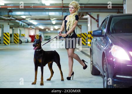 MOSCOW - JUN 01, 2015: Girl (with model release) with long blond hair in dress with the Doberman on leash in the underground parking Stock Photo