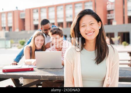 On foreground an asian schoolgirl staring front behind her multiracial friends. Young chinese teenage female student, smiling and looking at camera sitting with her classmates at university campus. High quality photo Stock Photo