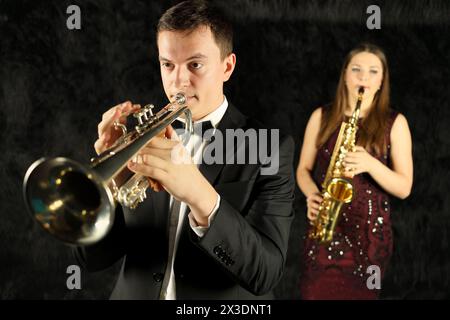 Man playing on trumpet in front of a woman with saxophone in a black room Stock Photo