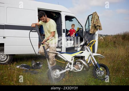 Man washes motorbike after training outdoor near minibus, little son looks at him from driver cabin. Stock Photo