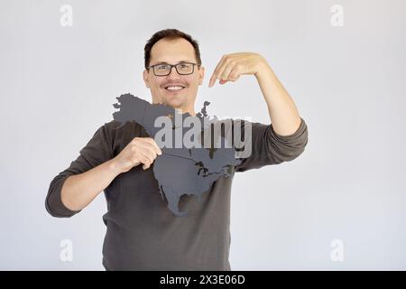 Smiling man in glasses holds puzzle map of Northern America and points finger at it. Stock Photo