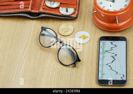 Glasses, smartphone with financial charts, wallet, crypto-coins, lying orange alarmclock on wooden desk. Stock Photo