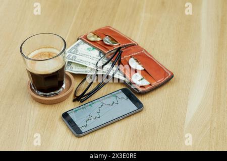 Glass of coffee, smartphone with financial charts, glasses and red wallet with coins and banknotes on wooden desk. Stock Photo