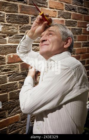 Elderly man stands near wall of red bricks with screwdriver in hand. Stock Photo