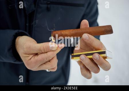 Close-up hands of man in jacket lighting tip of cigar with burning match. Stock Photo