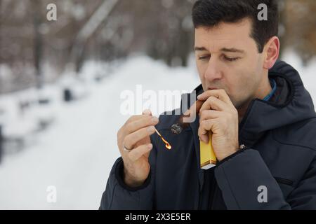 Man lights and puffs at cigar in winter snowy park. Stock Photo