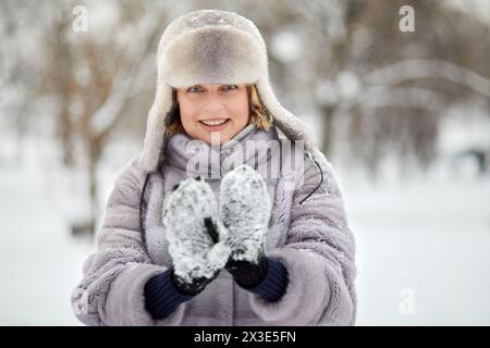 Smiling woman in fur coat and hat shows mittens with stuck snow in winter park. Stock Photo