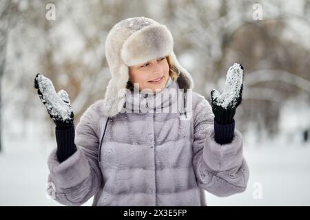 Smiling woman in fur coat and hat stands looking at mittens on her hands with stuck snow in winter park. Stock Photo