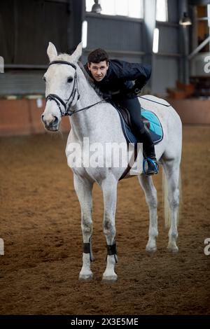 Man sits on horseback, bending over to mane at covered riding arena. Stock Photo