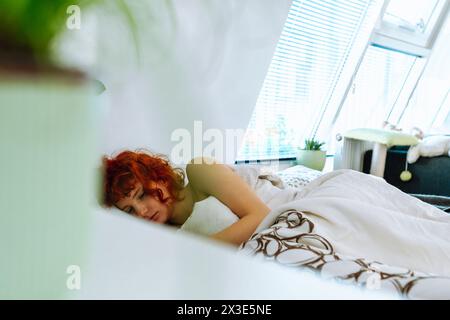 young red-haired woman lies in bed under blanket, uses smartphone, morning vacation concept Stock Photo
