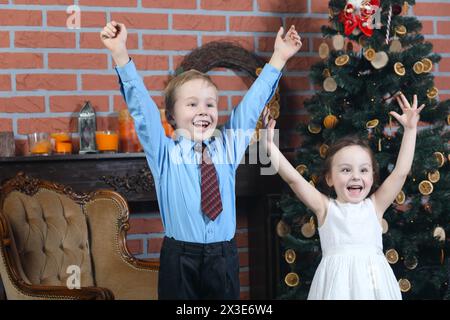 Boy and girl have fun with raised arms near christmas tree with dried oranges in brick studio, focus on boy Stock Photo