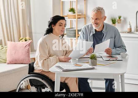 A disabled woman in a wheelchair and her husband sit at a table covered with papers, engaged in a thoughtful discussion. Stock Photo